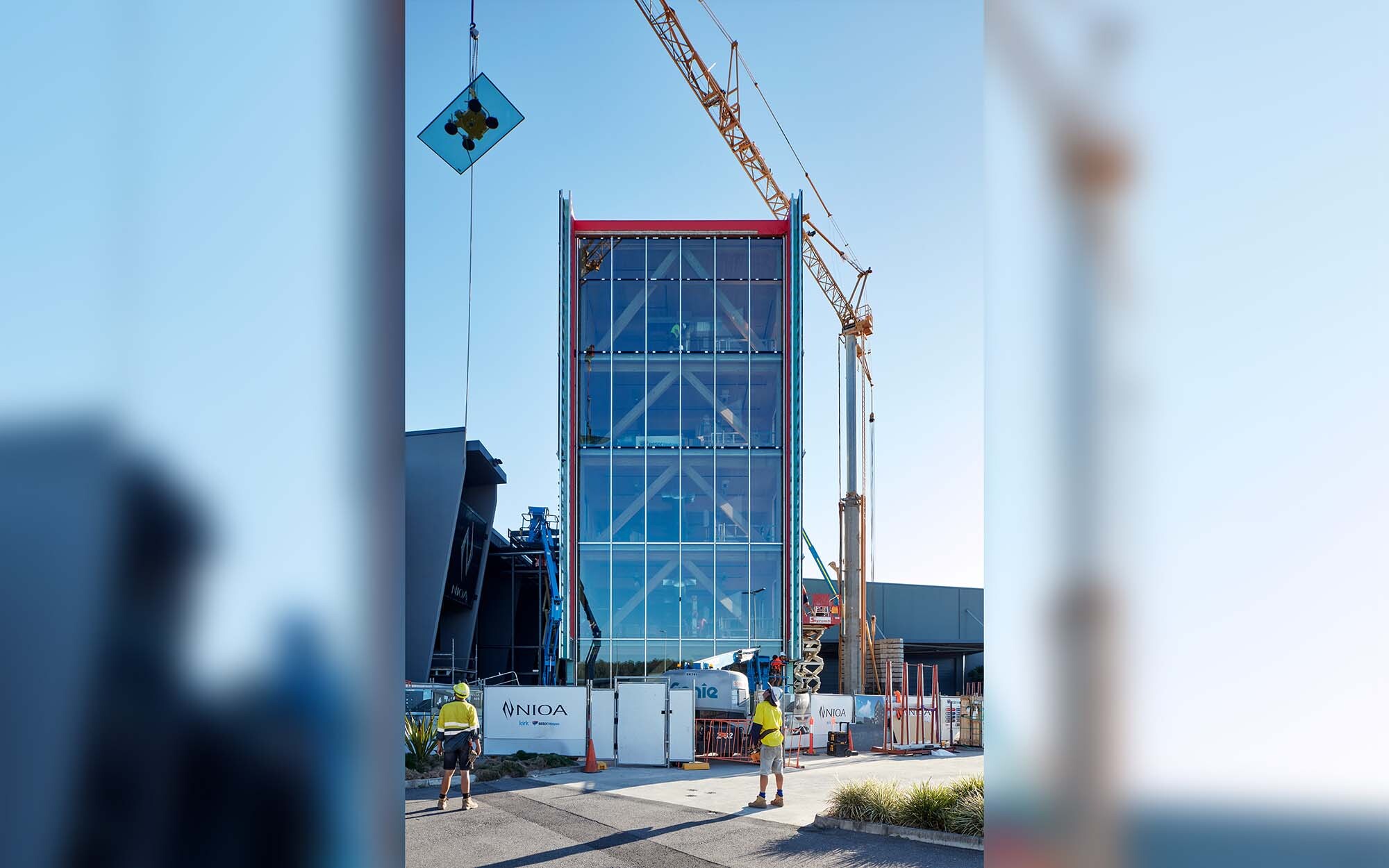 exterior view of NIOA Timber Tower under construction. Large yellow crane hoists glazing panel above structure.