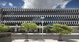 Long exterior elevation view of commercial architecture building designed by KIRK. Street perspective view shows geometric side facade and cladding under blue sky with clouds. Street runs parallel to building with green leafy trees on both sides of street.