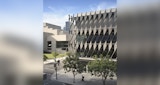 Corner street perspective of Kirk Studio designed commercial building on corner of street with buildings behind. Woman walks on sidewalk out front below evenly spaced trees. Exterior of building has geometric sun shading on the exterior.