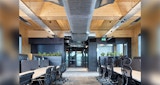 Alternate view of interior open concept workspace inside Kirk Studio designed Timber Tower for NIOA. two large parallel rows of desks with black rolling chairs. view to glass doors into exterior hall. Timber walls and ceiling left exposed with grey carpeting.